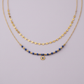 Collier double rang doré avec perles bleues et pendentif soleil sur fond lilas
